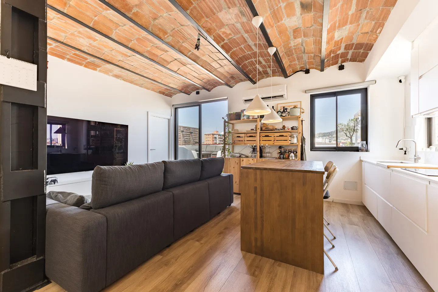 Modern living room with a vaulted brick ceiling, dark grey sofa, wooden kitchen island, and white cabinets.