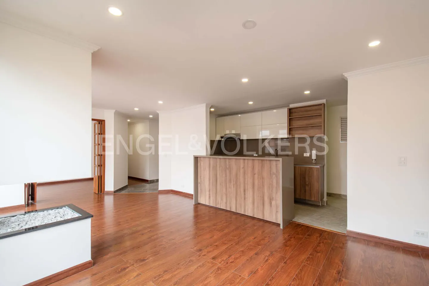 Open-concept living space with hardwood floors, white walls, and recessed lighting. A kitchen with a wood-paneled island is visible.