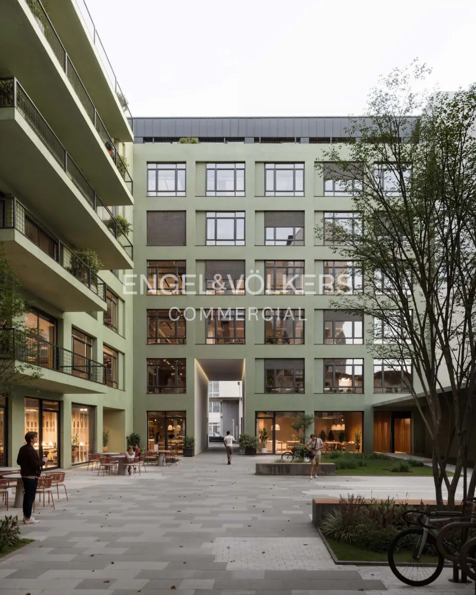 Exterior view of a light green commercial building with a courtyard, tables, and people. The Engel & Völkers logo is visible on the building.
