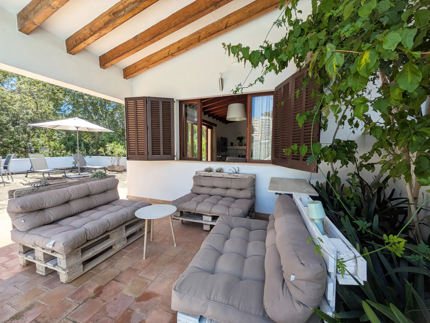 Outdoor patio with pallet furniture. Cushioned pallet sofas and a small white table sit on a brick patio. Brown shutters frame a window.