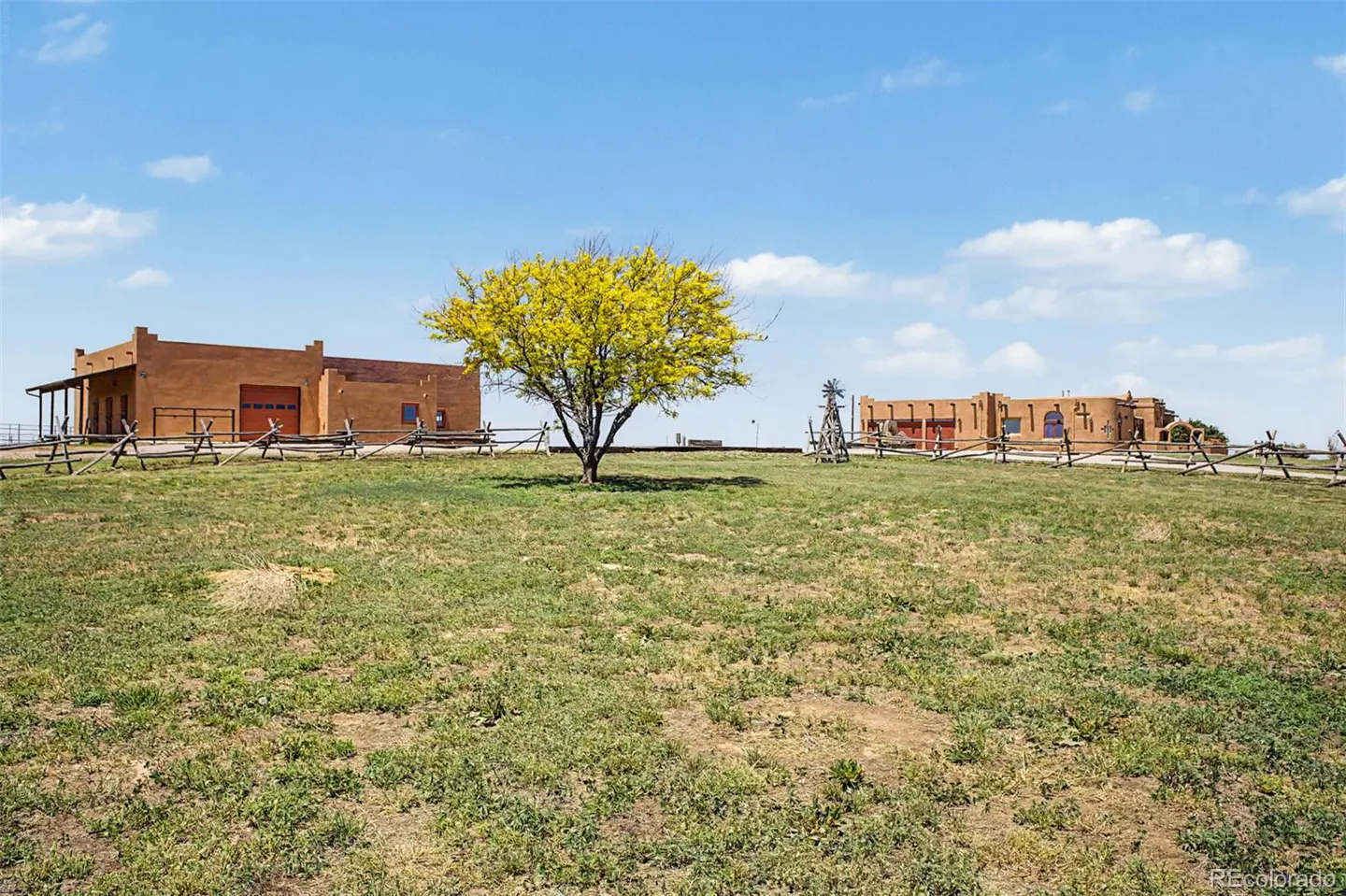 Two adobe-style buildings sit on a grassy field under a blue sky, separated by a vibrant yellow tree and wooden fencing.