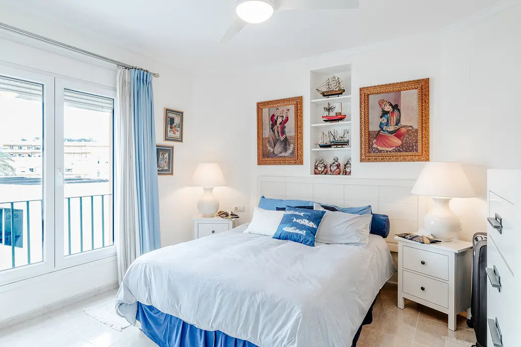 Bright bedroom with white walls, a bed with blue accents, and nautical decor. A window with blue curtains overlooks a balcony.