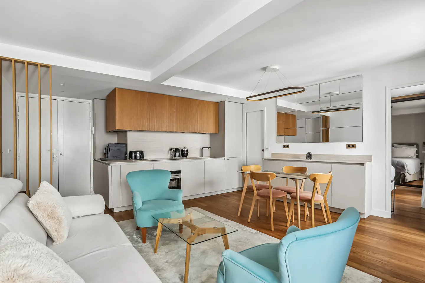 Bright, modern apartment interior with white sofa, blue chairs, wood floors, and a kitchen with wood cabinets.