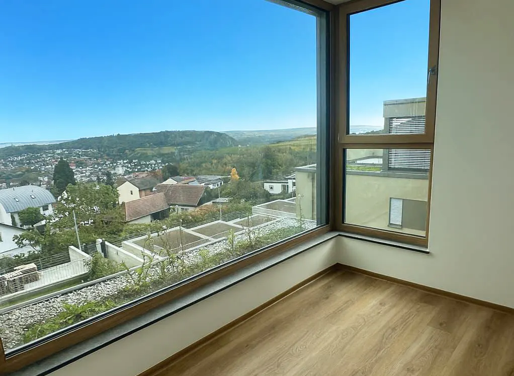 A corner of a room with wood floors and a large window showing a town and green hills under a blue sky.