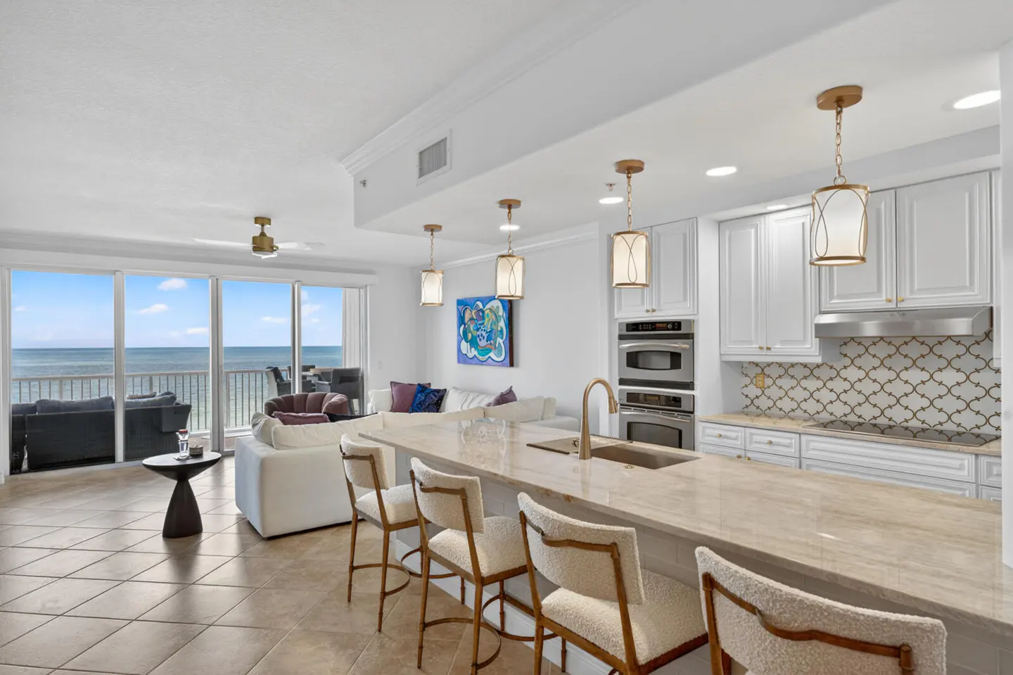 Modern open-concept kitchen with white cabinets, stone island, and bar stools, overlooking a living area and ocean view.