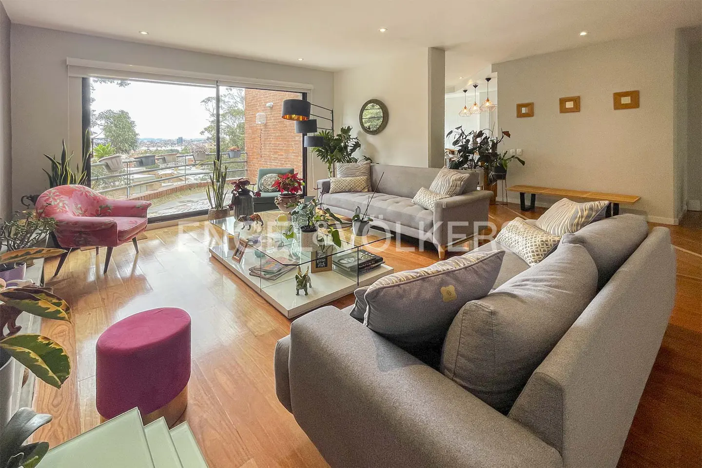 Bright living room with hardwood floors, gray sofas, pink chair, and a large window overlooking a cityscape.