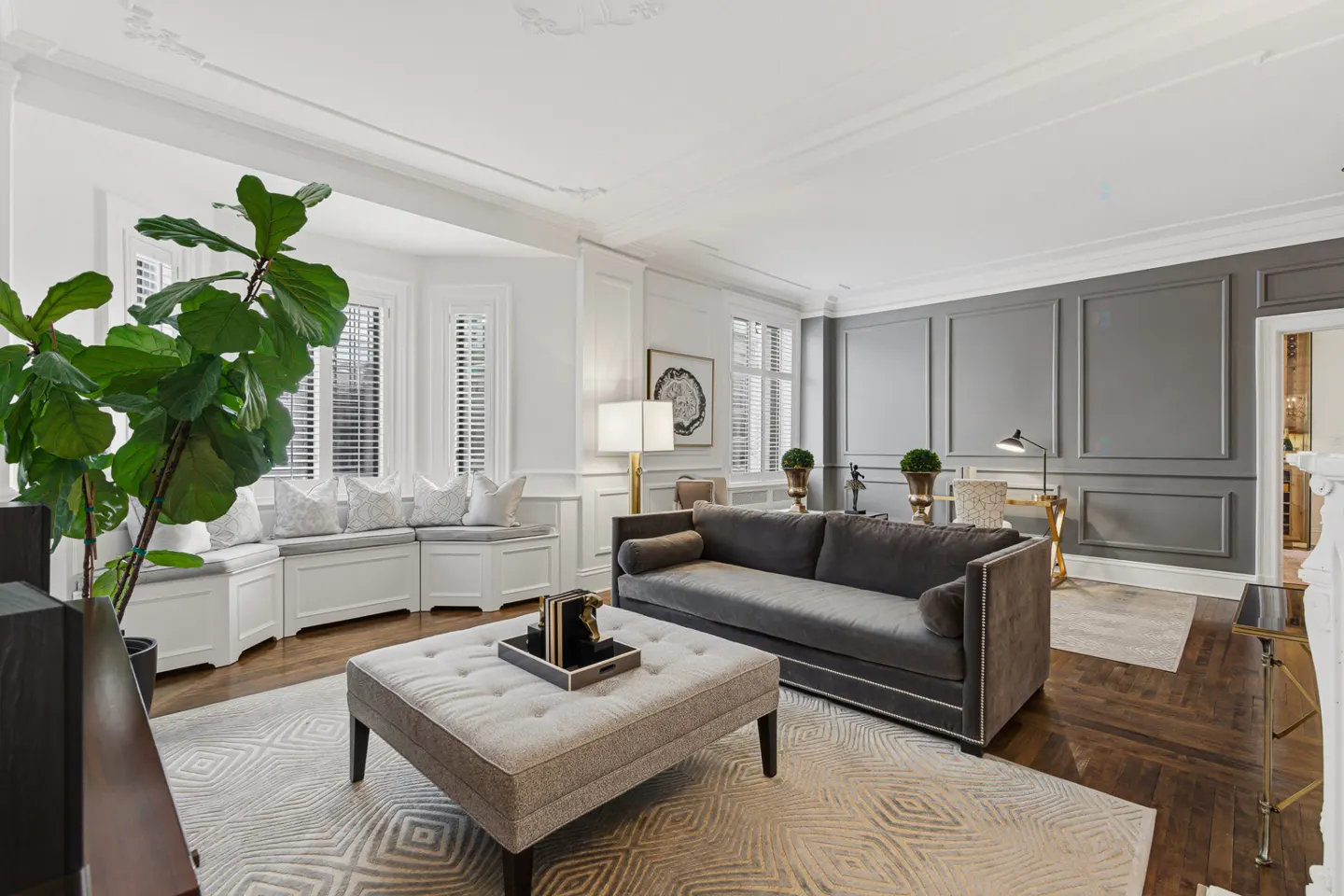 Living room with gray sofa, ottoman, and gray paneled wall. A large green plant sits near a window seat.