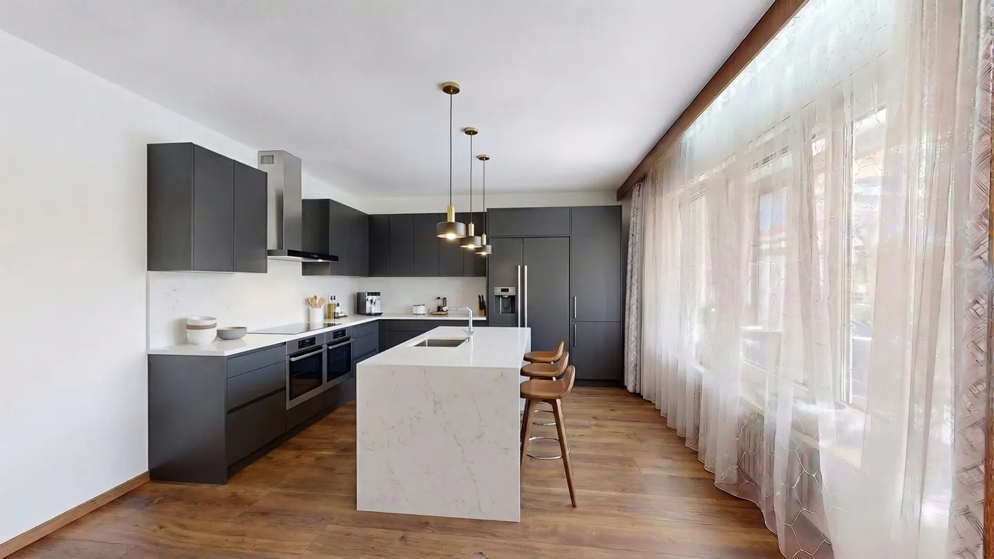 A modern kitchen with gray cabinets, a white marble island with three stools, and sheer white curtains.