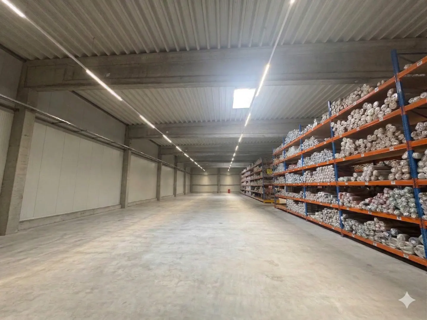 A wide shot of a large, empty warehouse with concrete floors and a high ceiling with fluorescent lighting. Shelves filled with rolled goods line one wall.