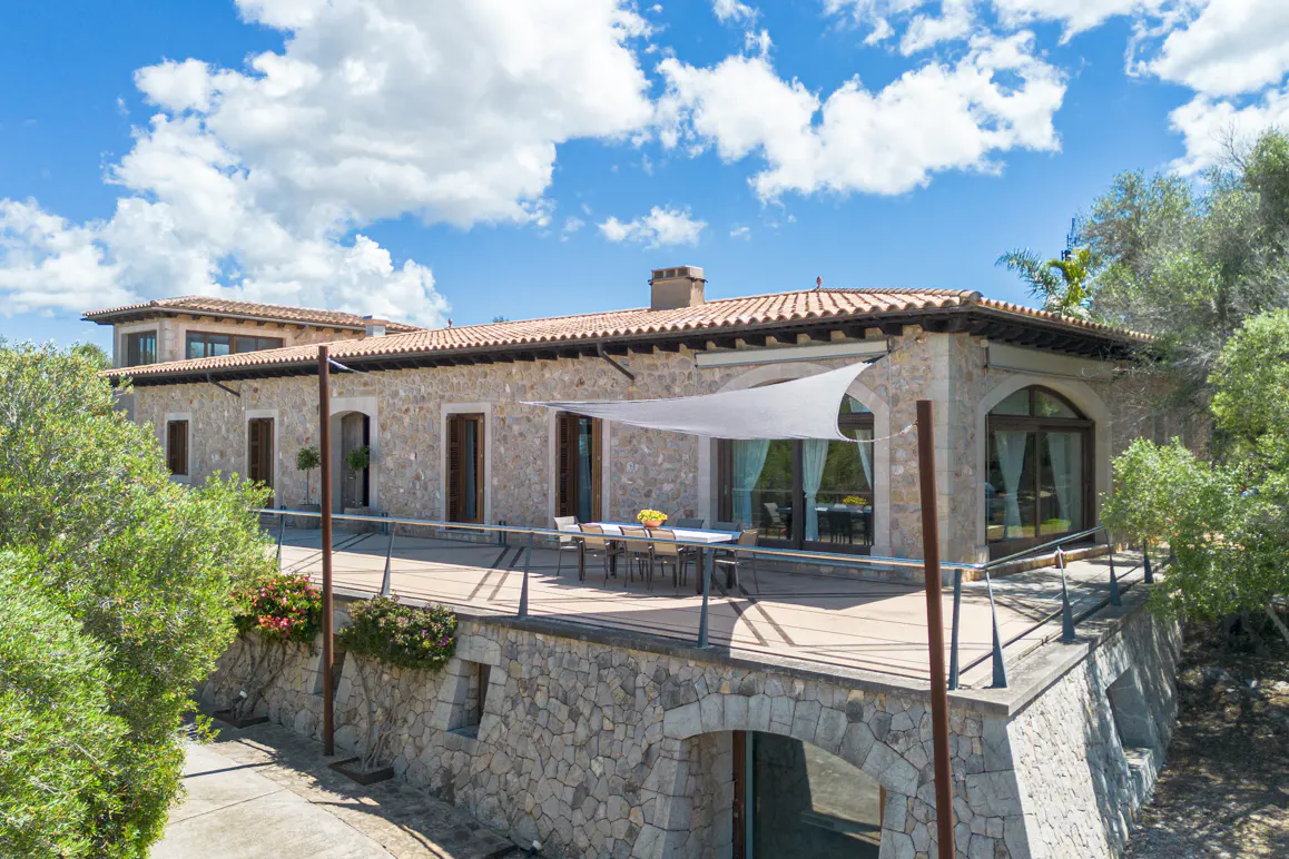 Stone house with a red tile roof and a patio with a table and chairs under a white sunshade.