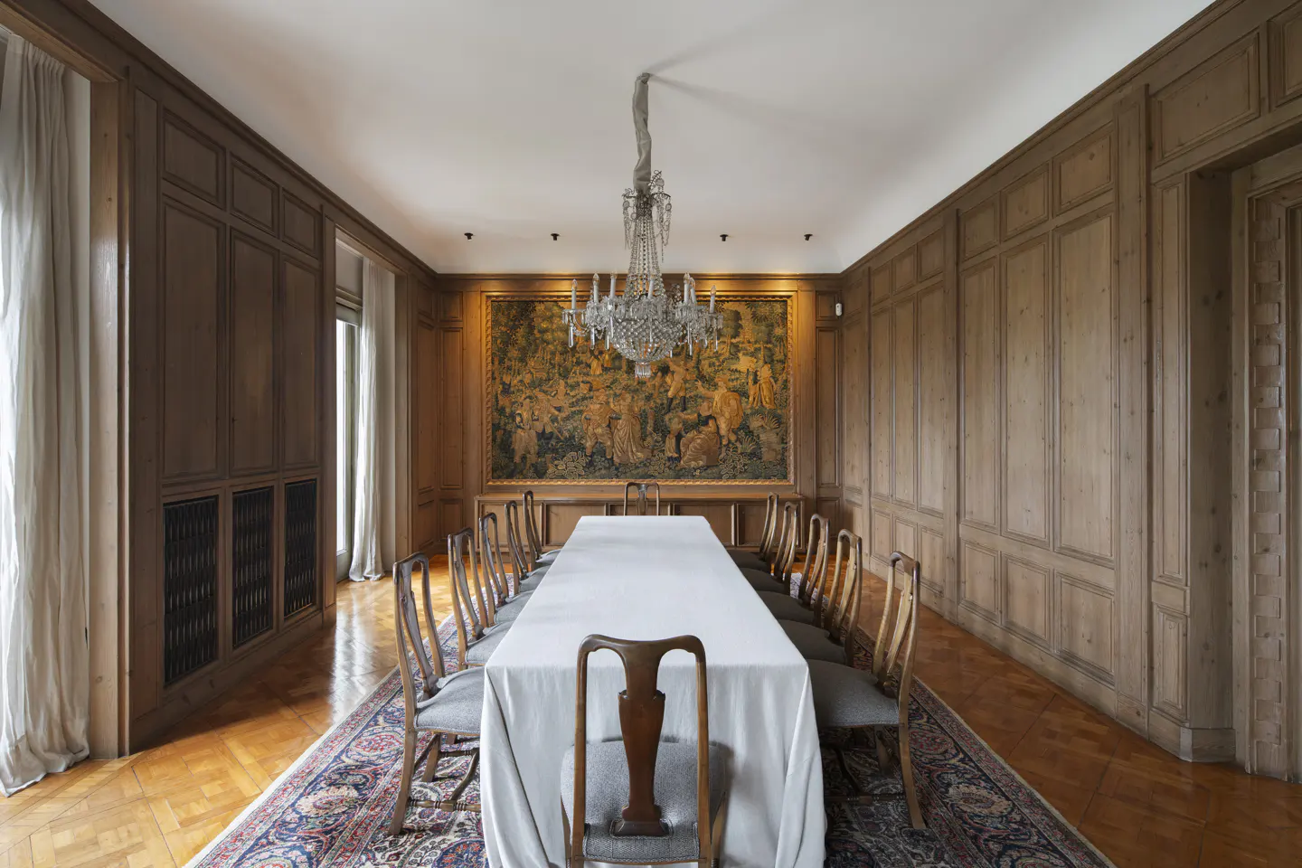 Formal dining room with wood-paneled walls, a long table with a white cloth, crystal chandelier, and a large wall tapestry.