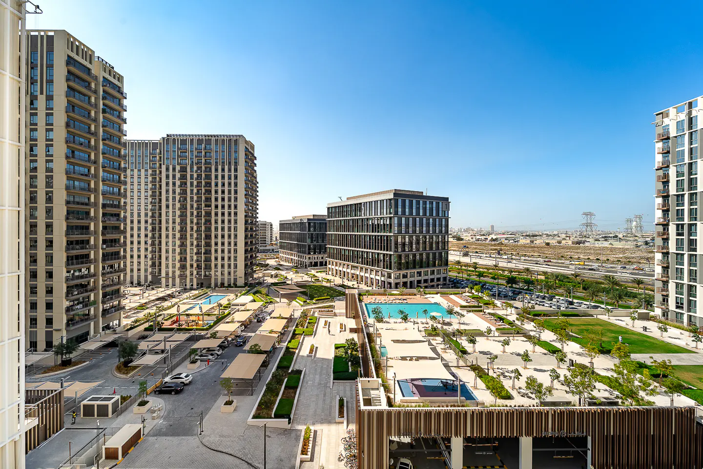 View of modern apartment buildings with pools, parking, and landscaping under a clear blue sky.
