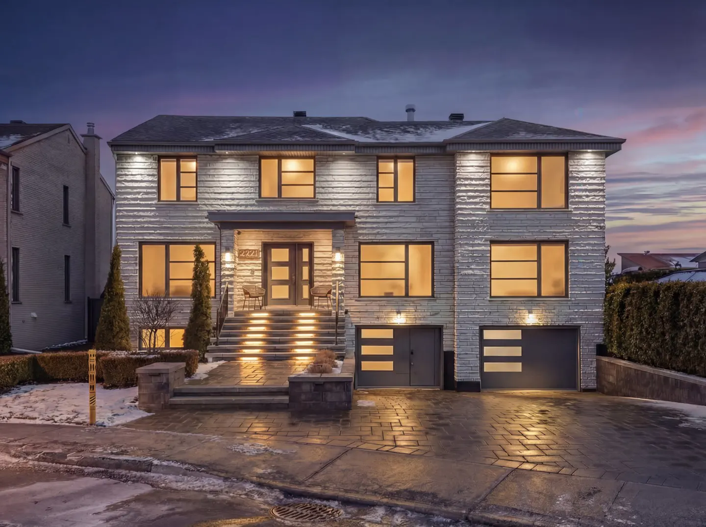 Exterior view of a two-story house with gray brick, a paved driveway, and illuminated steps leading to the front door.