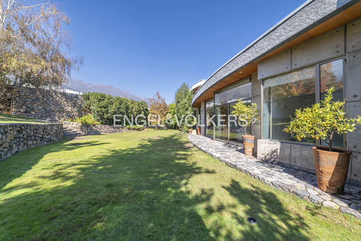 Lush green lawn with stone wall and modern house. Potted citrus trees line the stone path. Blue sky and mountain backdrop.