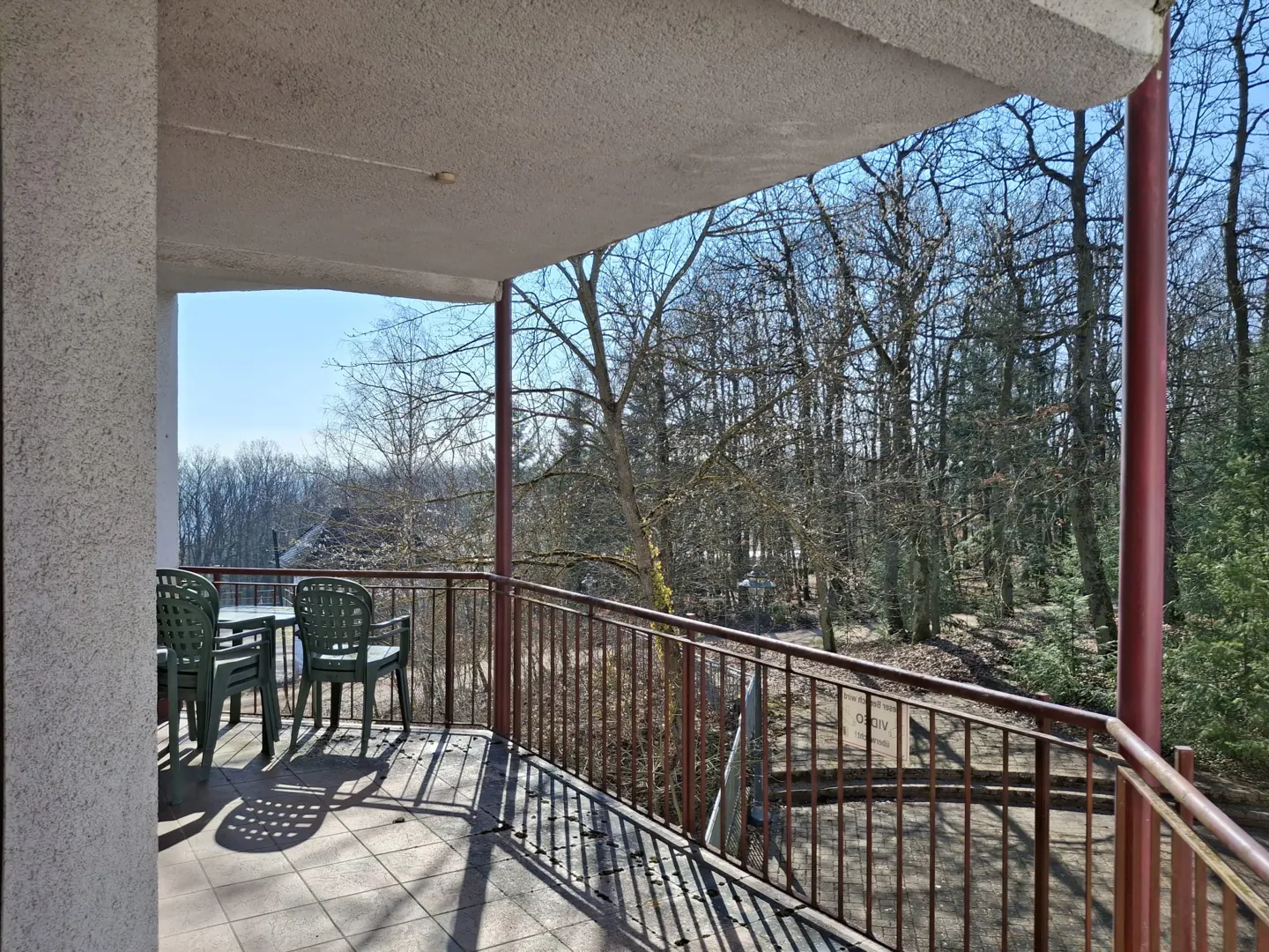 Balcony view with green chairs and table, brown railing, and a forest backdrop under a blue sky.