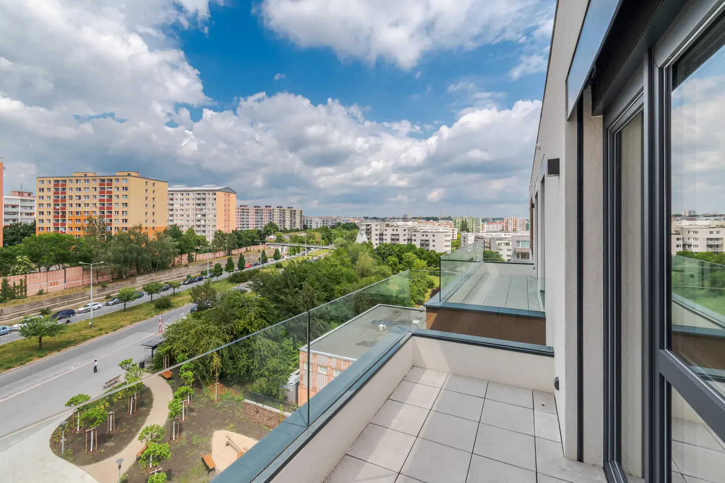 Modern apartment balcony with glass railings overlooking a city street, green trees, and residential buildings under a sky.
