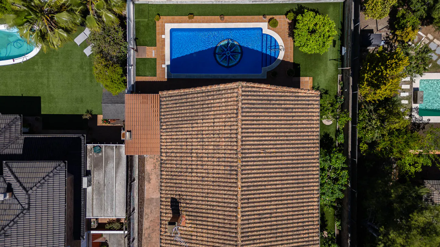 Aerial view of a house with a brown tiled roof and a rectangular pool with a compass design in the center. Lush green lawns and trees surround the property.