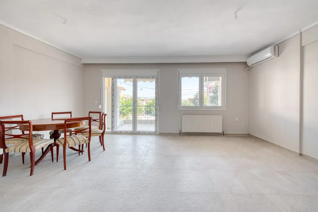 Bright, empty room with a round wooden table and chairs, white walls, and a sliding glass door to a balcony.