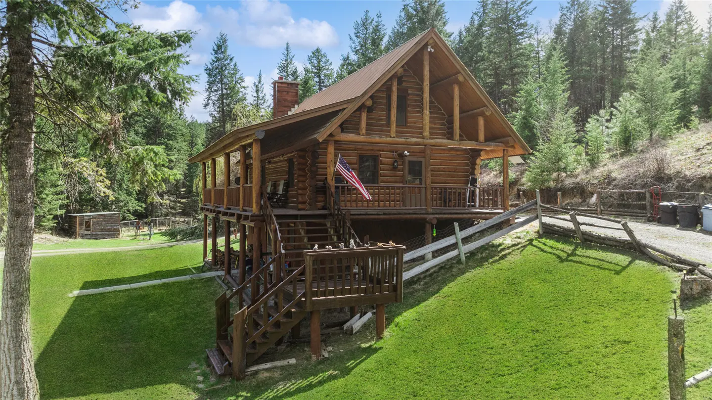 Two-story log cabin with a brown roof, chimney, and American flag on a green lawn with trees in the background.