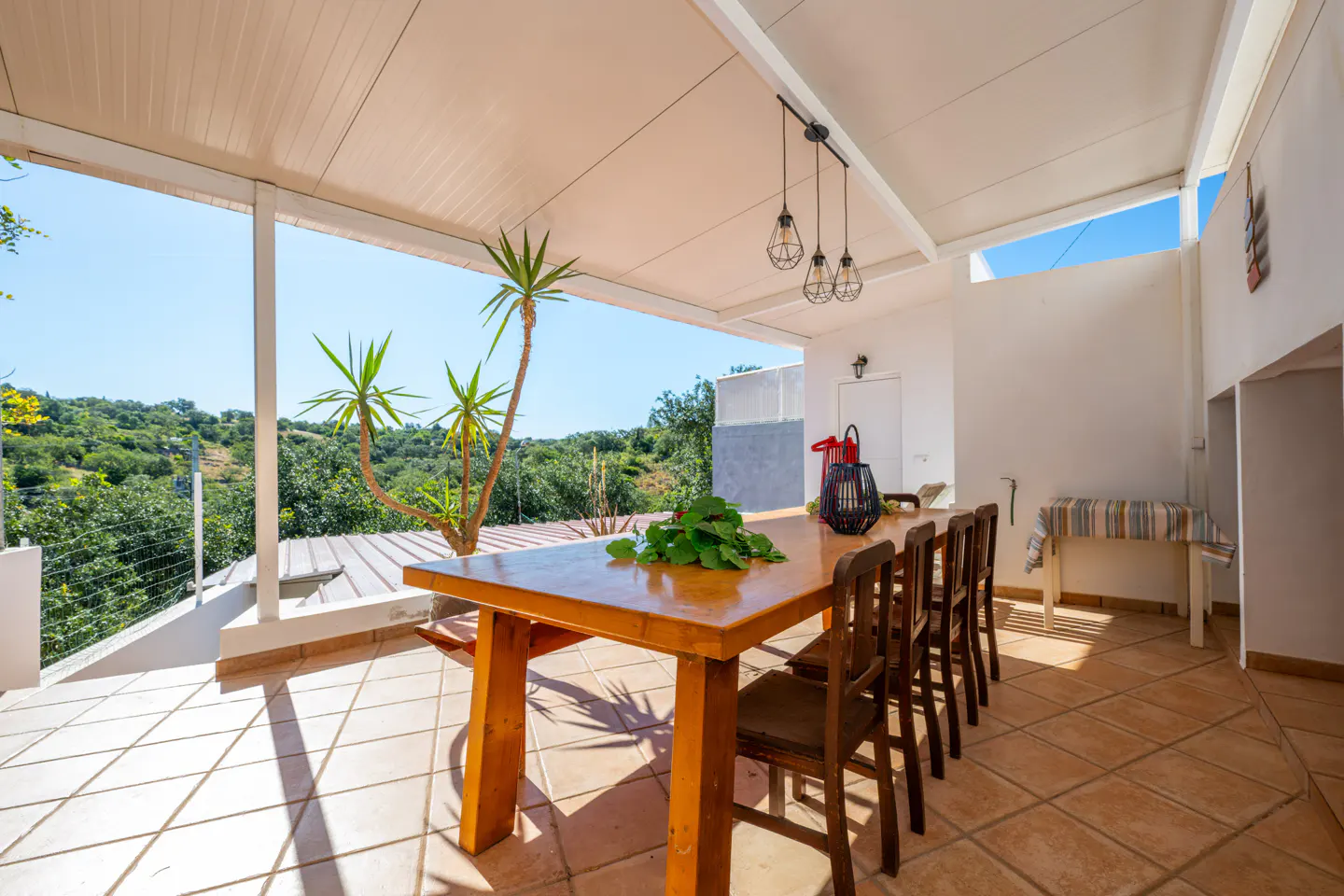 Covered patio with a long wooden table, six chairs, and a view of green hills. Three pendant lights hang above the table.