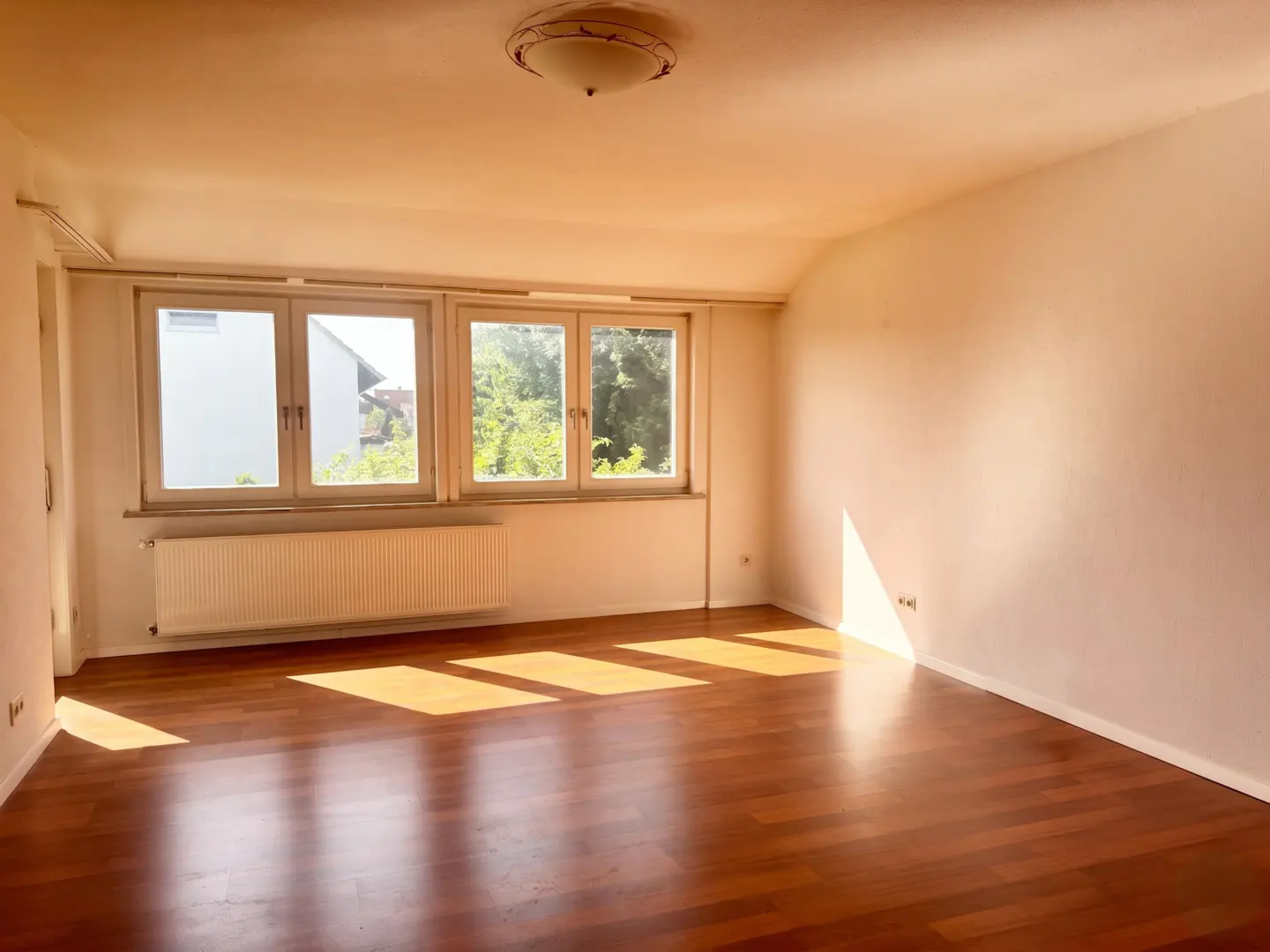 Bright, empty room with hardwood floors, white walls, and three windows letting in sunlight. A radiator sits beneath the windows.