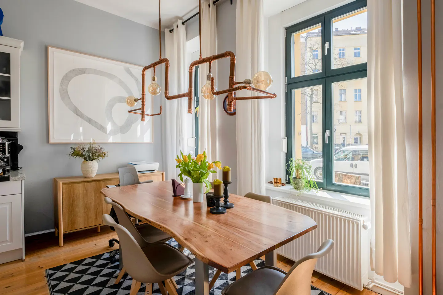 A dining room with a live edge wood table, modern chairs, and a copper pipe light fixture. A black and white rug sits under the table.