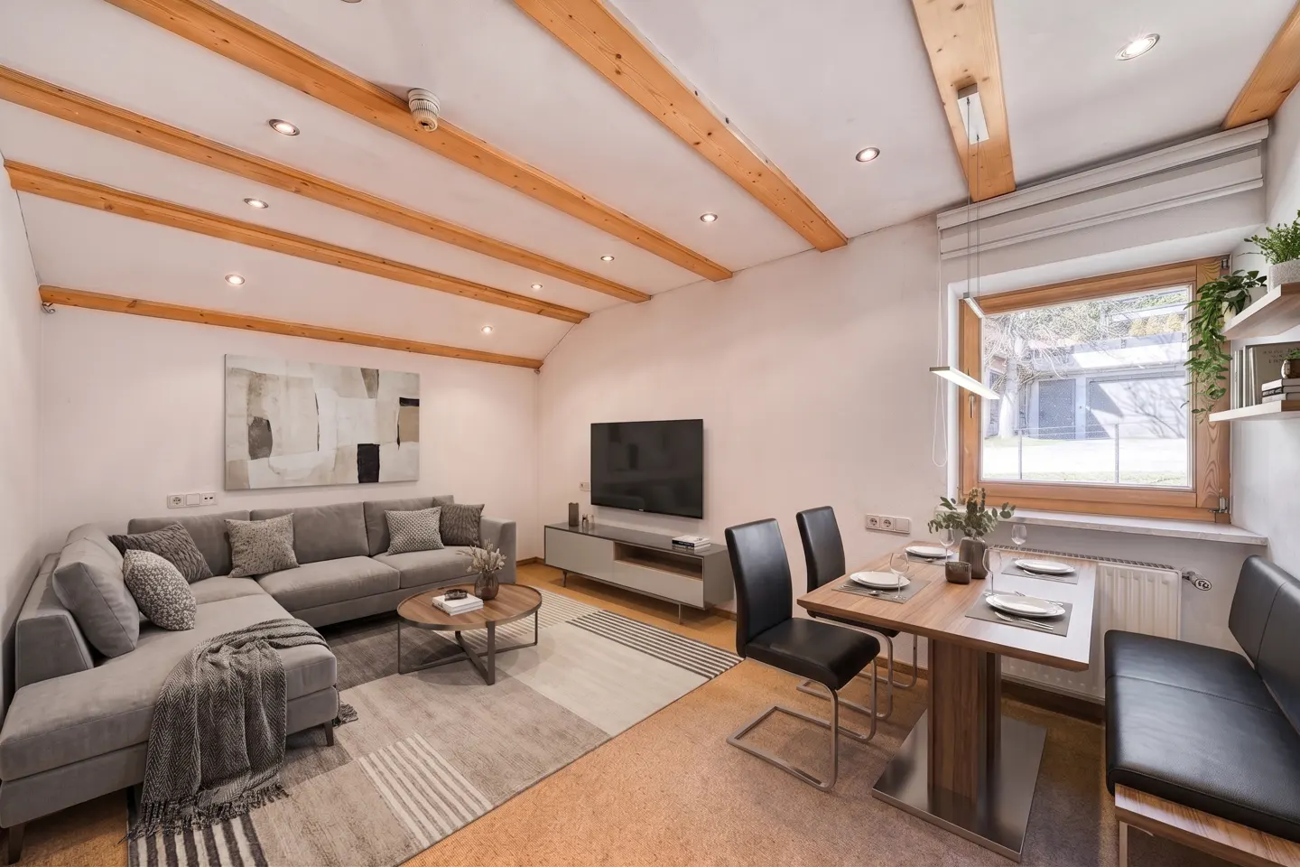 Living room with gray sectional sofa, wood beams, dining table, and large window. Neutral tones and modern decor.