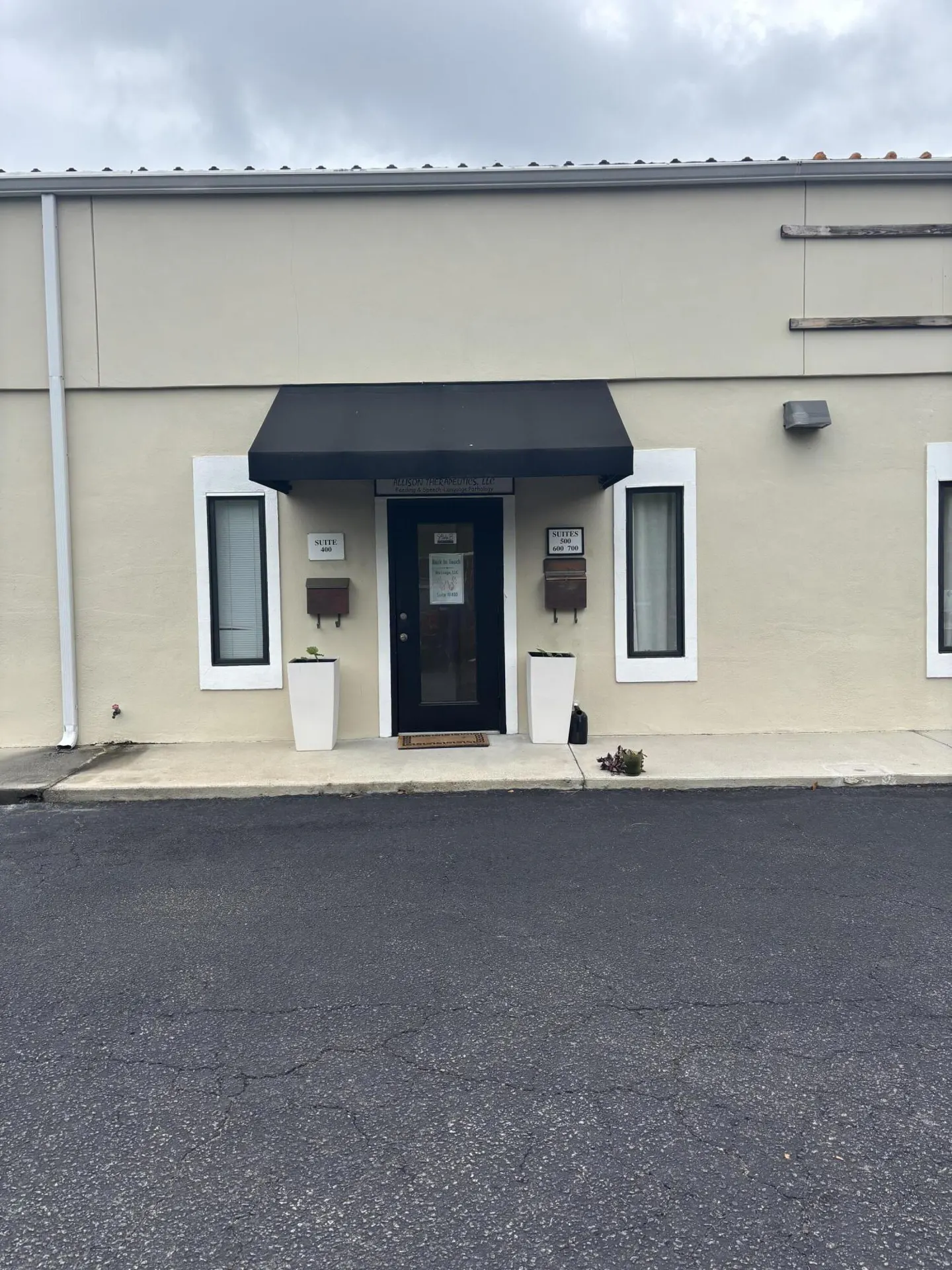 Tan building with a black awning over the entrance. White planters flank the door, and windows are on either side. Asphalt parking lot in front.