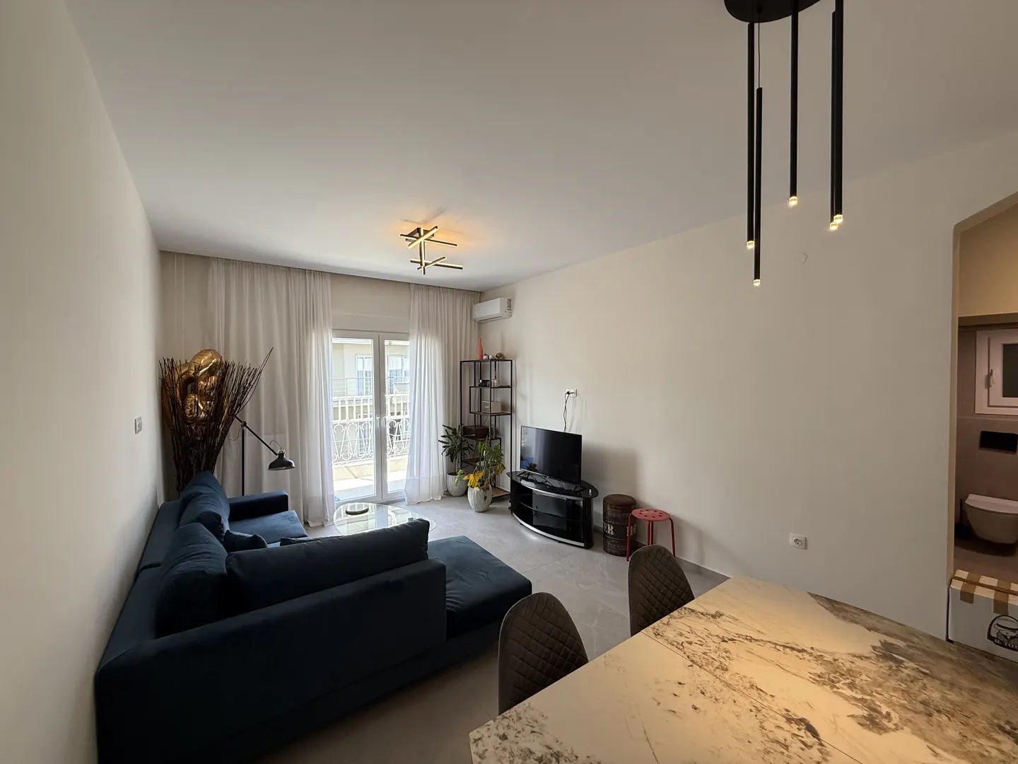 Bright living room with a blue sofa, TV, marble table, and balcony with white curtains.