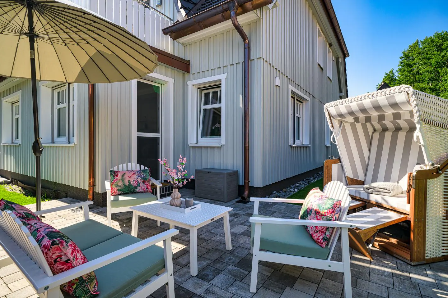 Outdoor patio with white furniture, floral pillows, and a striped beach chair. The patio is paved with gray stones. A light gray house is in the background.