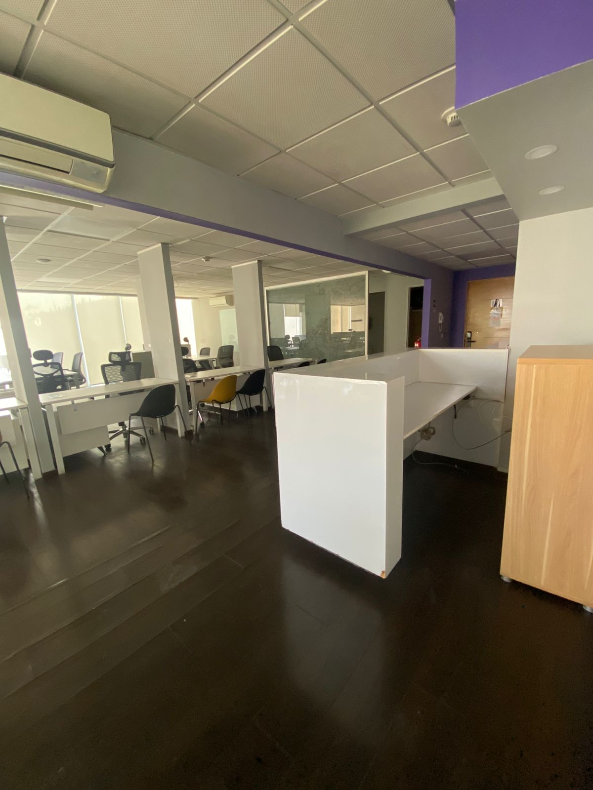 A modern office space with white desks, black chairs, and a polished dark floor. The ceiling is white with a purple accent wall in the background.