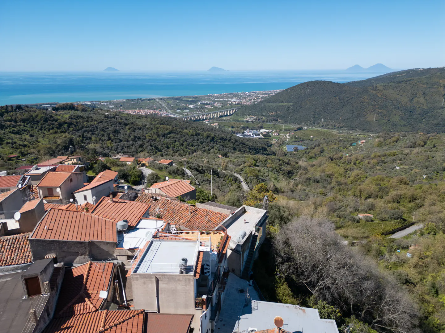 Scenic view of a village with red-tiled roofs, green hills, and the blue sea with islands in the distance under a clear sky.
