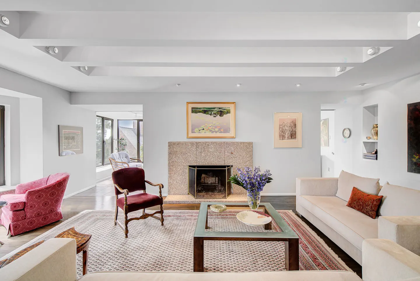 Bright living room with white walls, recessed lighting, and hardwood floors. A stone fireplace is centered with art above. Beige sofas and patterned chairs furnish the space.