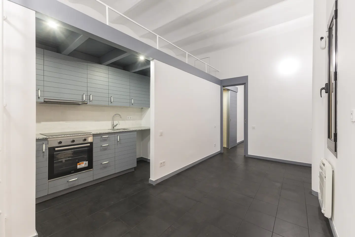 A modern apartment interior with gray cabinets, black tile floors, and white walls. A loft area is visible above the kitchen.