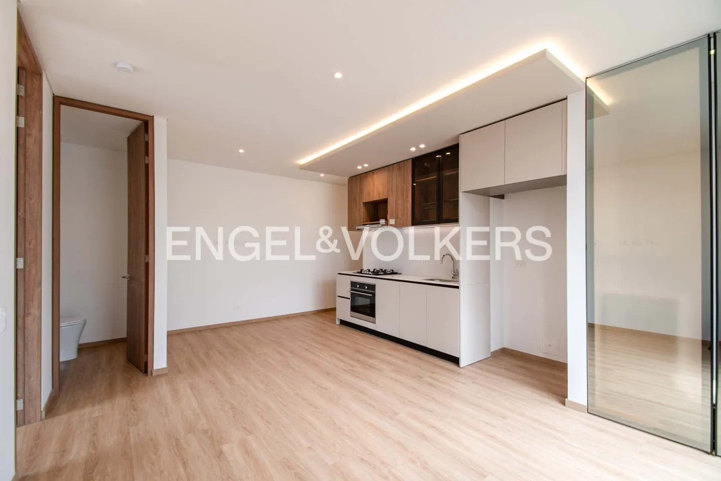 A bright, modern apartment interior with wood floors, white walls, and a compact kitchen with white cabinets.