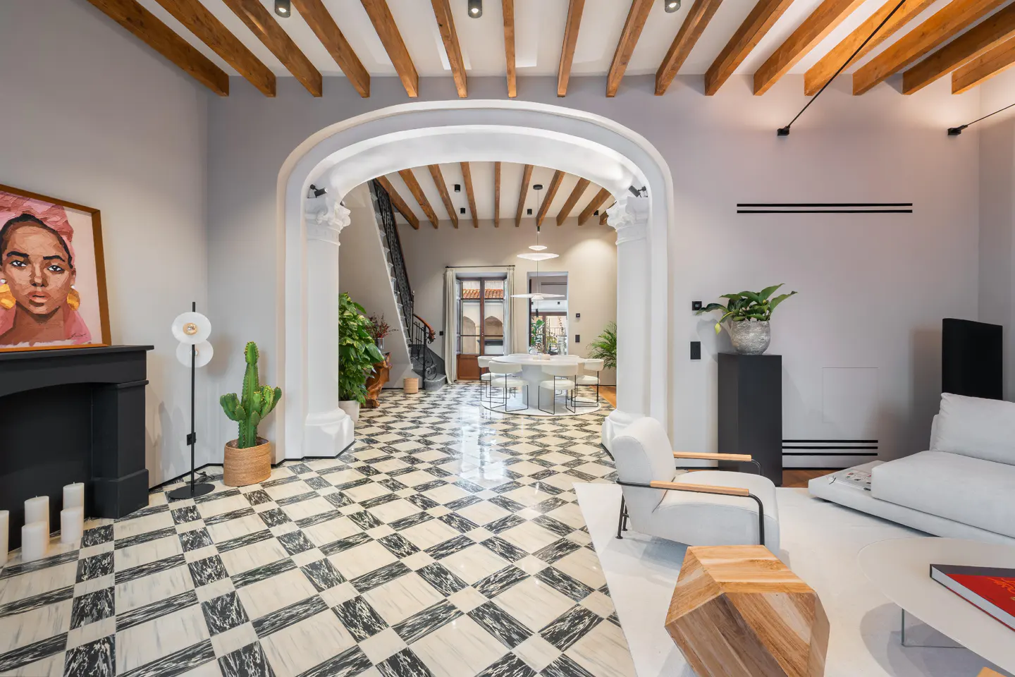 A bright, modern living room with black and white checkered floors, a white archway, and wooden ceiling beams. A black fireplace and white furniture add contrast.