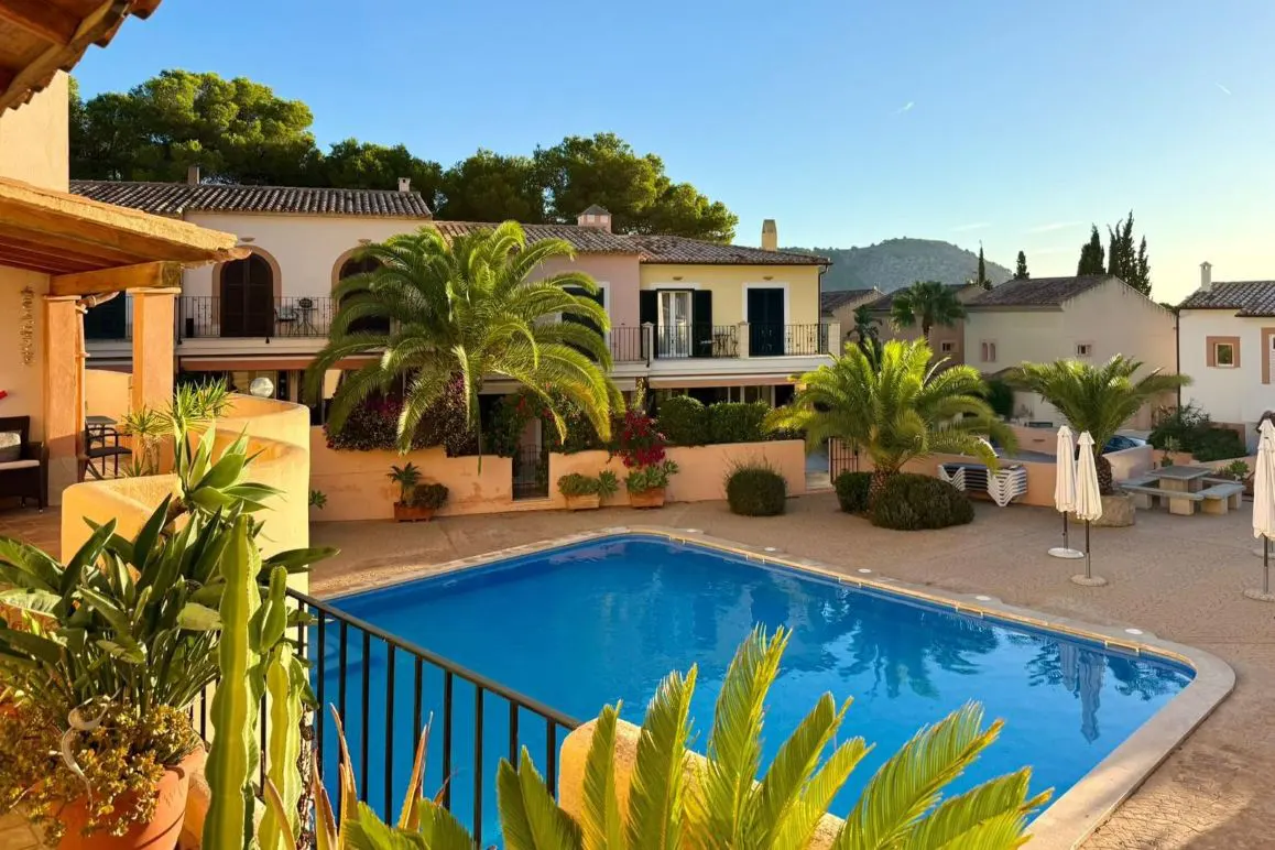 Sunny courtyard with a blue swimming pool, palm trees, and peach-colored buildings under a clear blue sky.