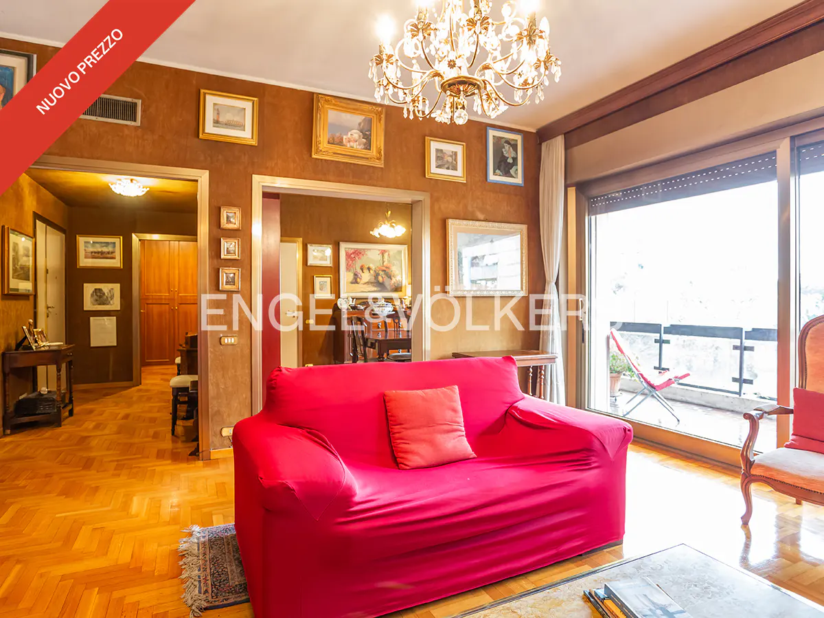 A living room with a red sofa, wood floors, and a chandelier. Paintings hang on the brown walls. A balcony is visible through sliding glass doors.