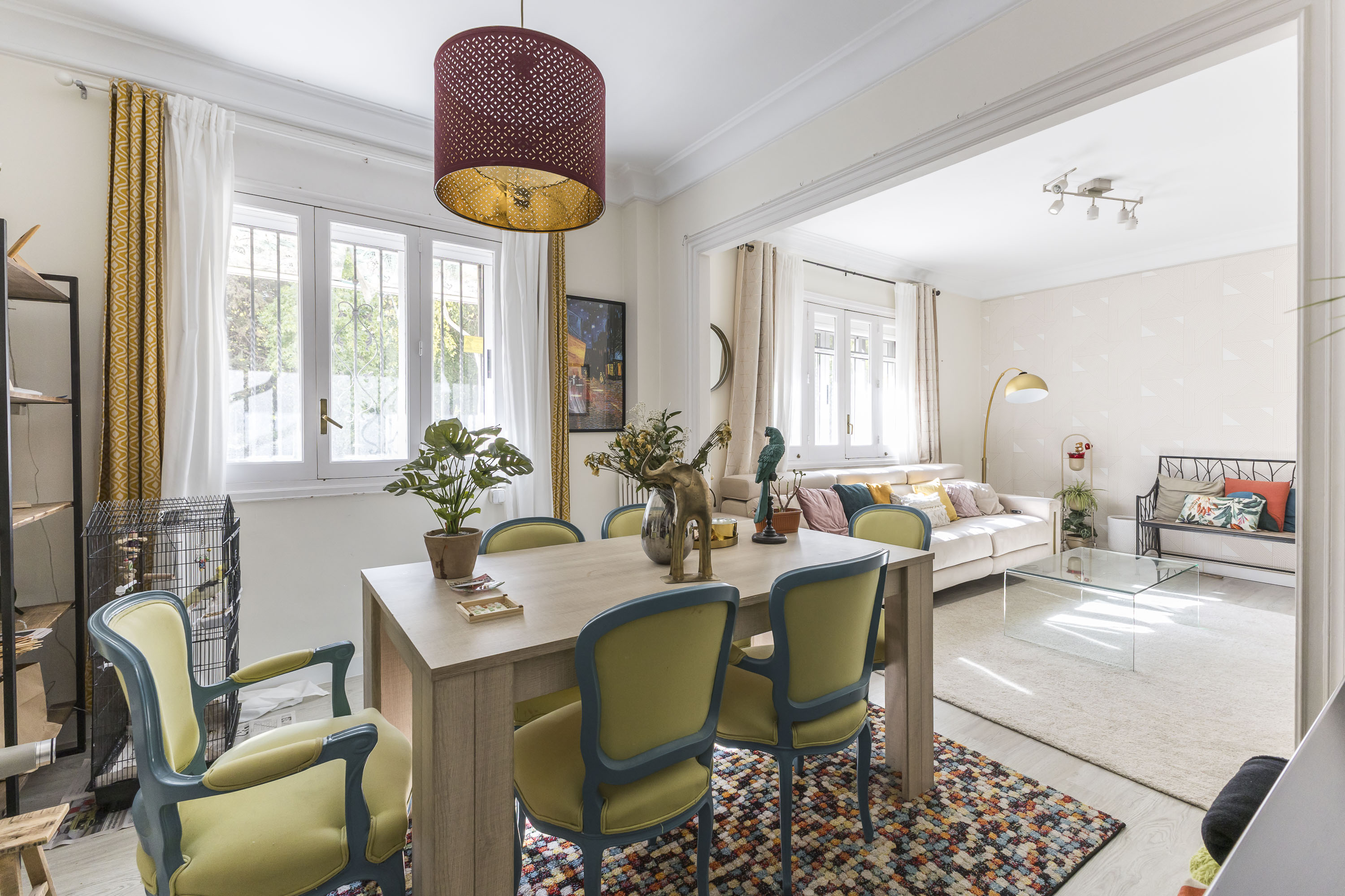 Dining area with a wooden table, lime green chairs, and a red pendant light, opening into a bright living room with a sofa. [AI-generated text]