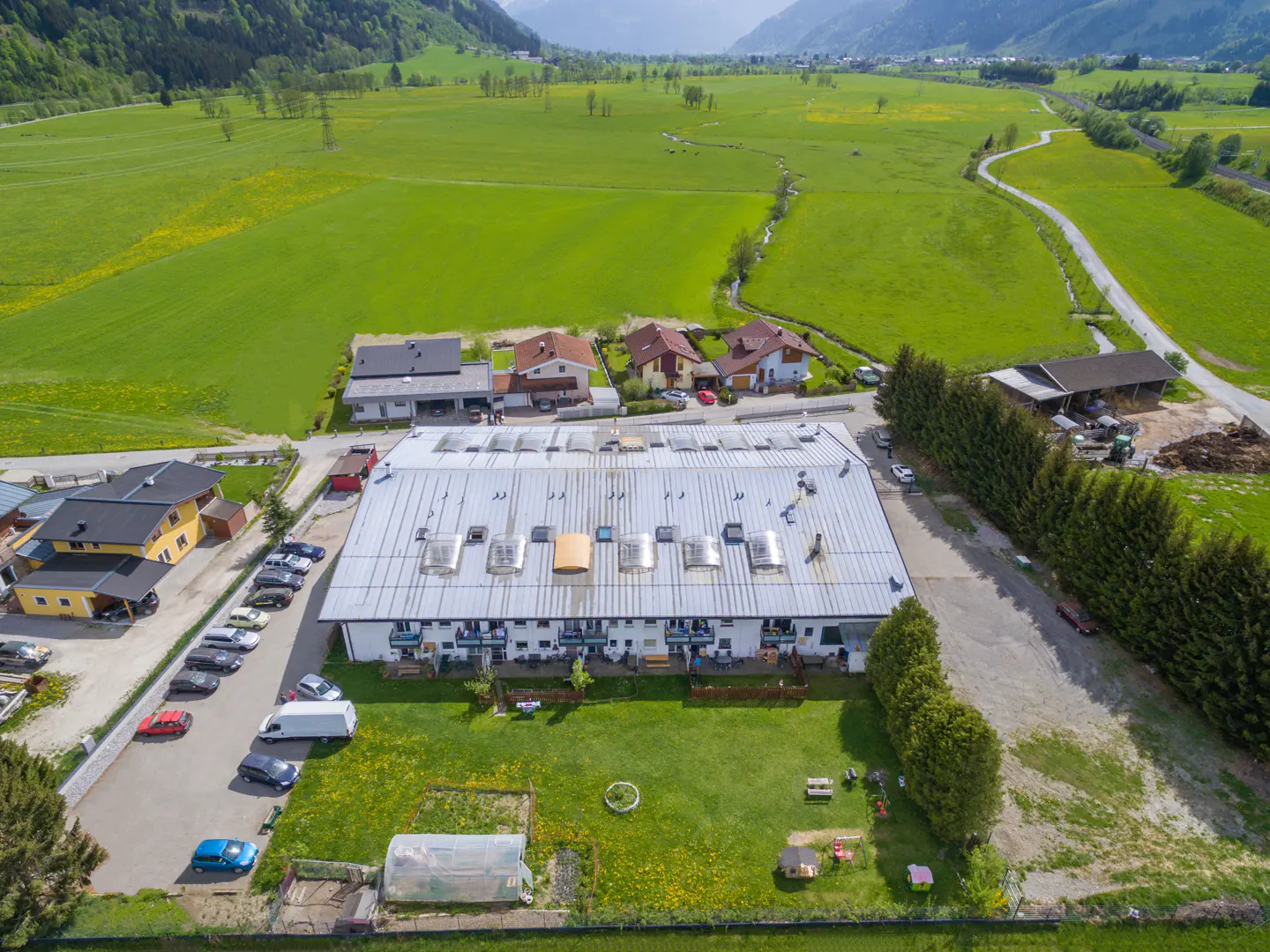Aerial view of a long, white apartment building with a silver roof and skylights, surrounded by green grass and fields.