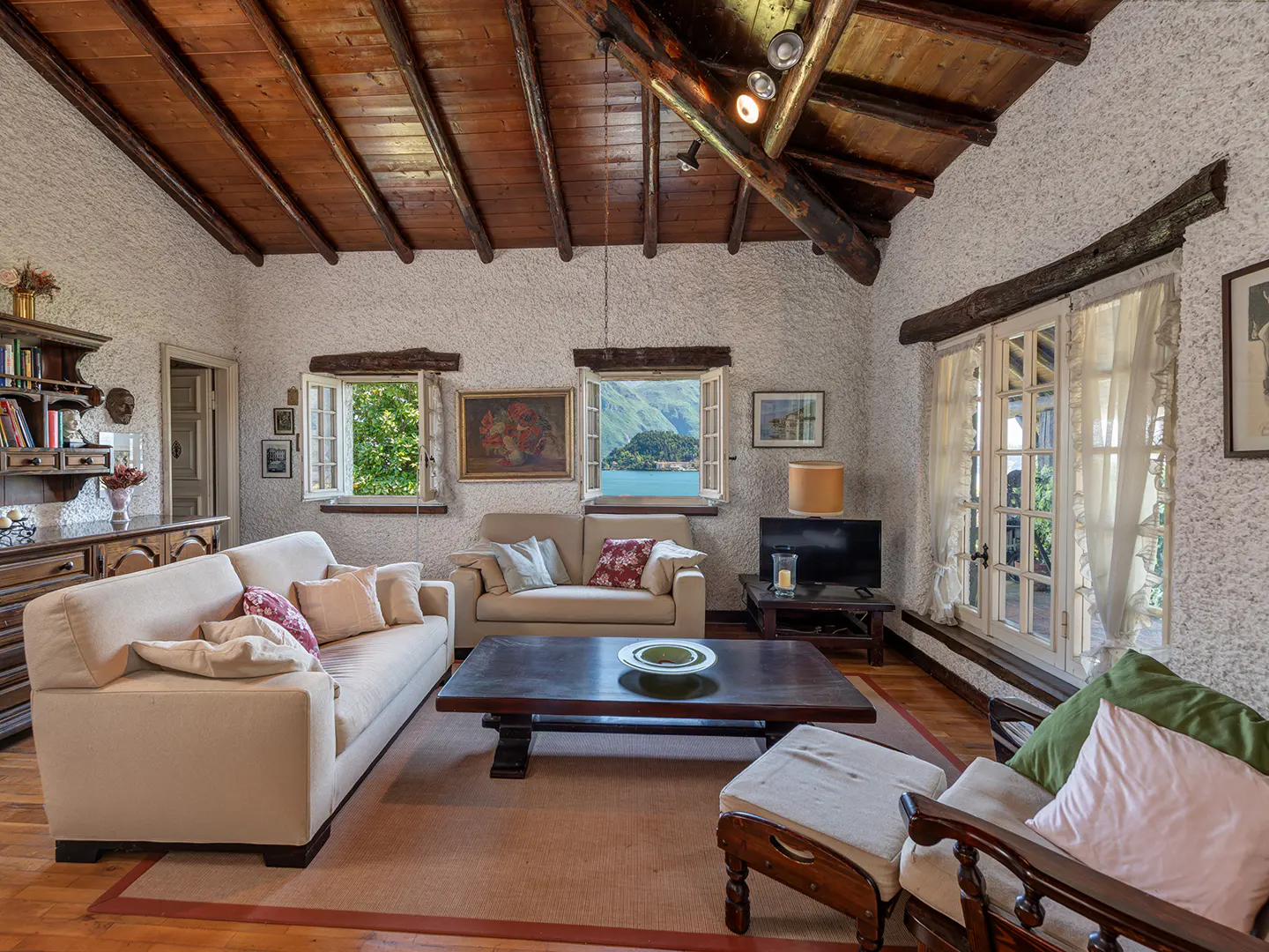 Living room with textured walls, wood ceiling, and large windows. Beige sofas, dark wood table, and a view of a lake.