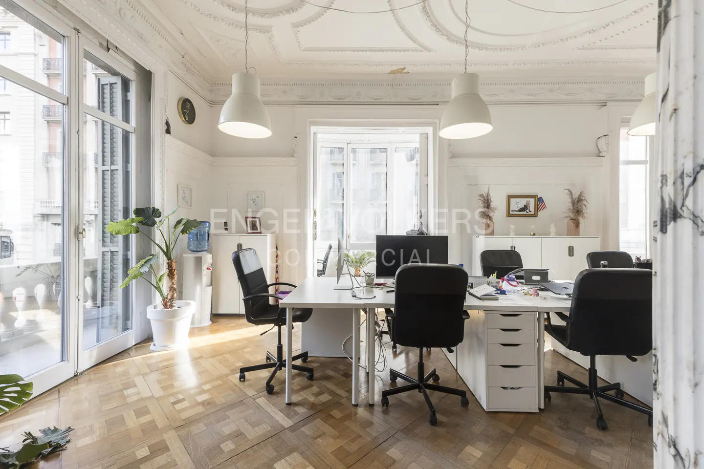 Bright office space with white walls, parquet floors, and large windows. White desks and black chairs are arranged for multiple workstations.