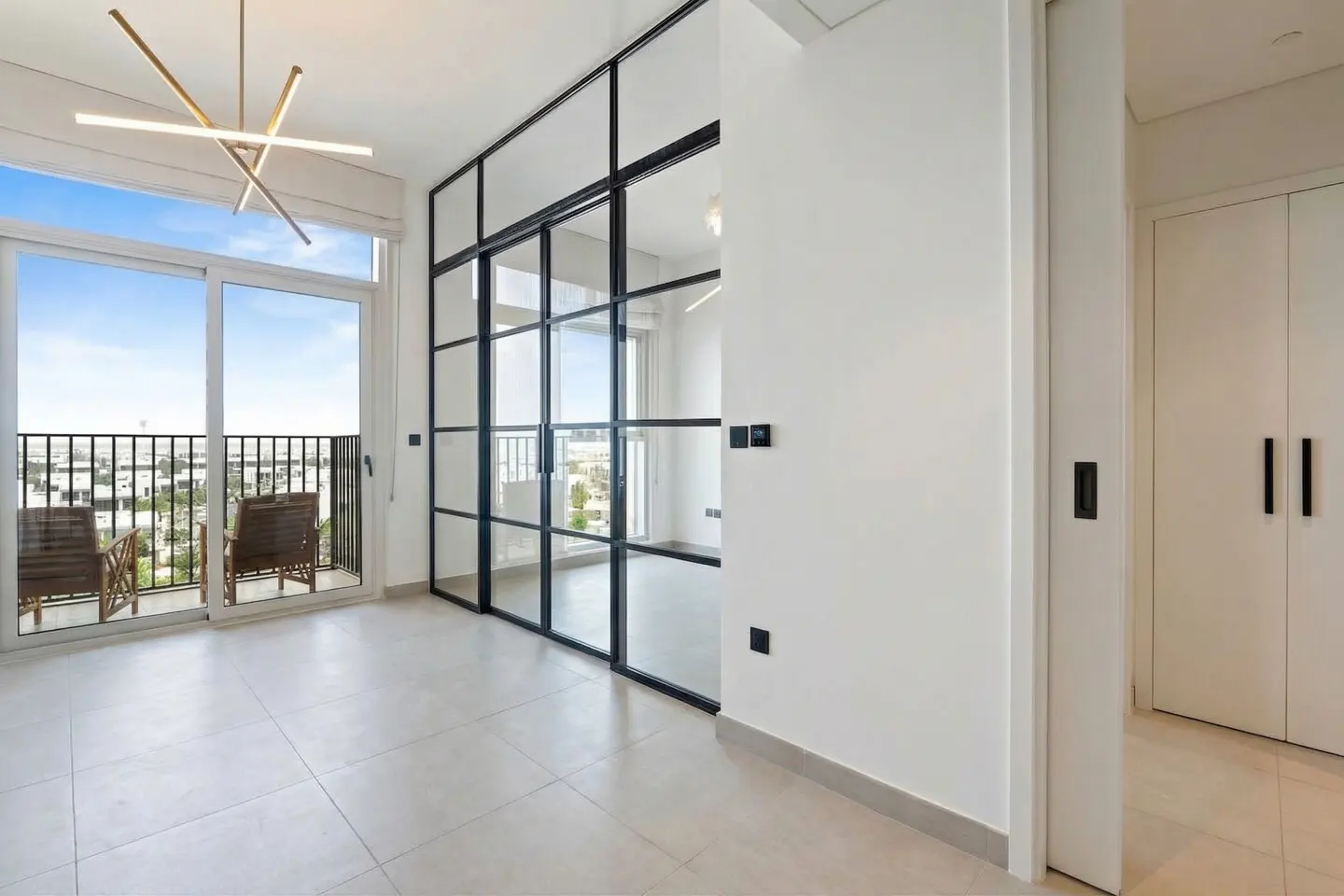 Bright, modern room with a balcony. Black framed glass wall, white walls, and tile floor. Two chairs on the balcony with a city view.