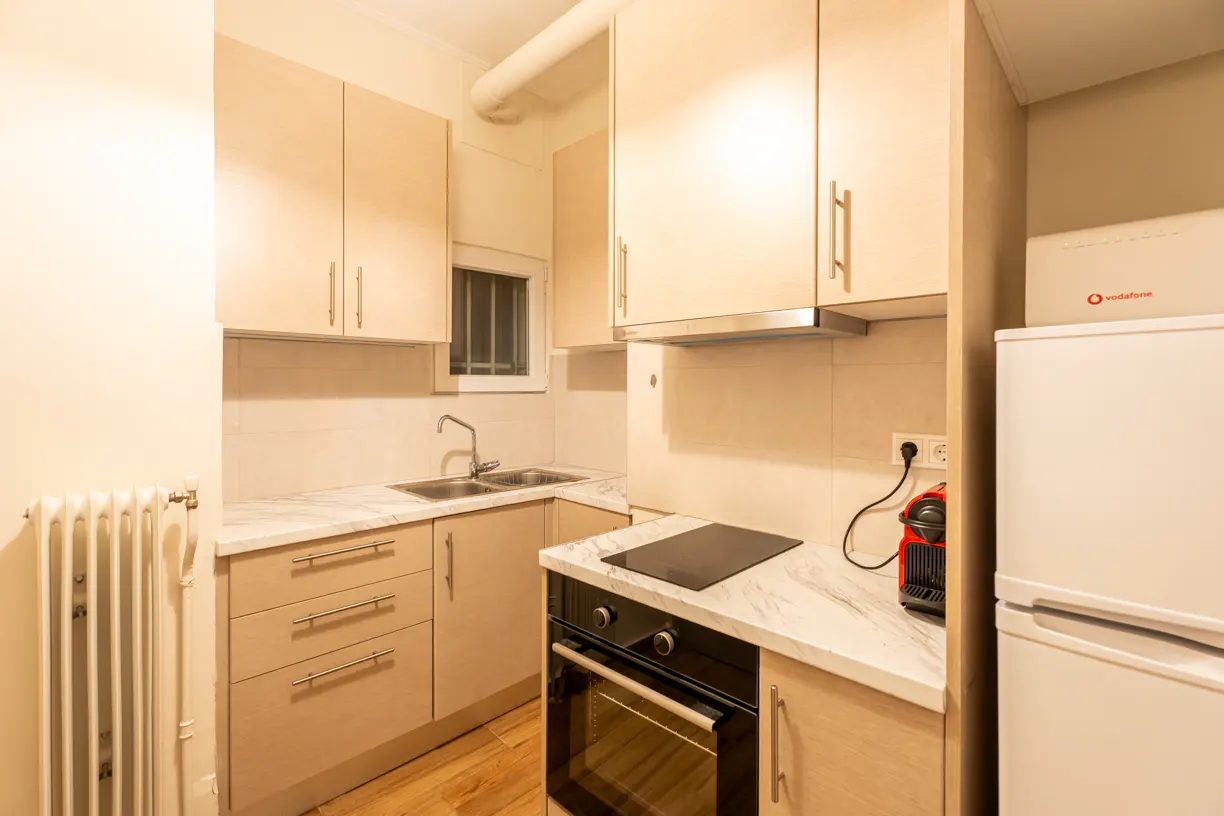 A compact kitchen with light wood cabinets, marble countertops, and a white refrigerator. A black stovetop and oven are visible.