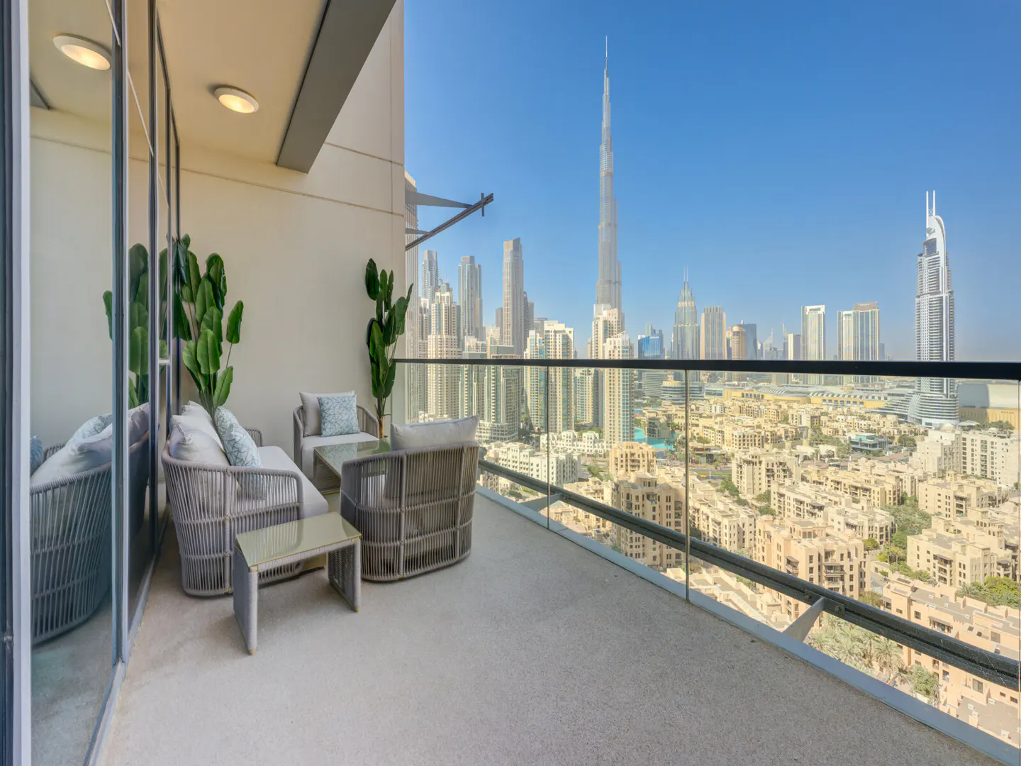 Balcony with gray wicker furniture, plants, and a glass railing overlooking the Dubai skyline on a clear day.