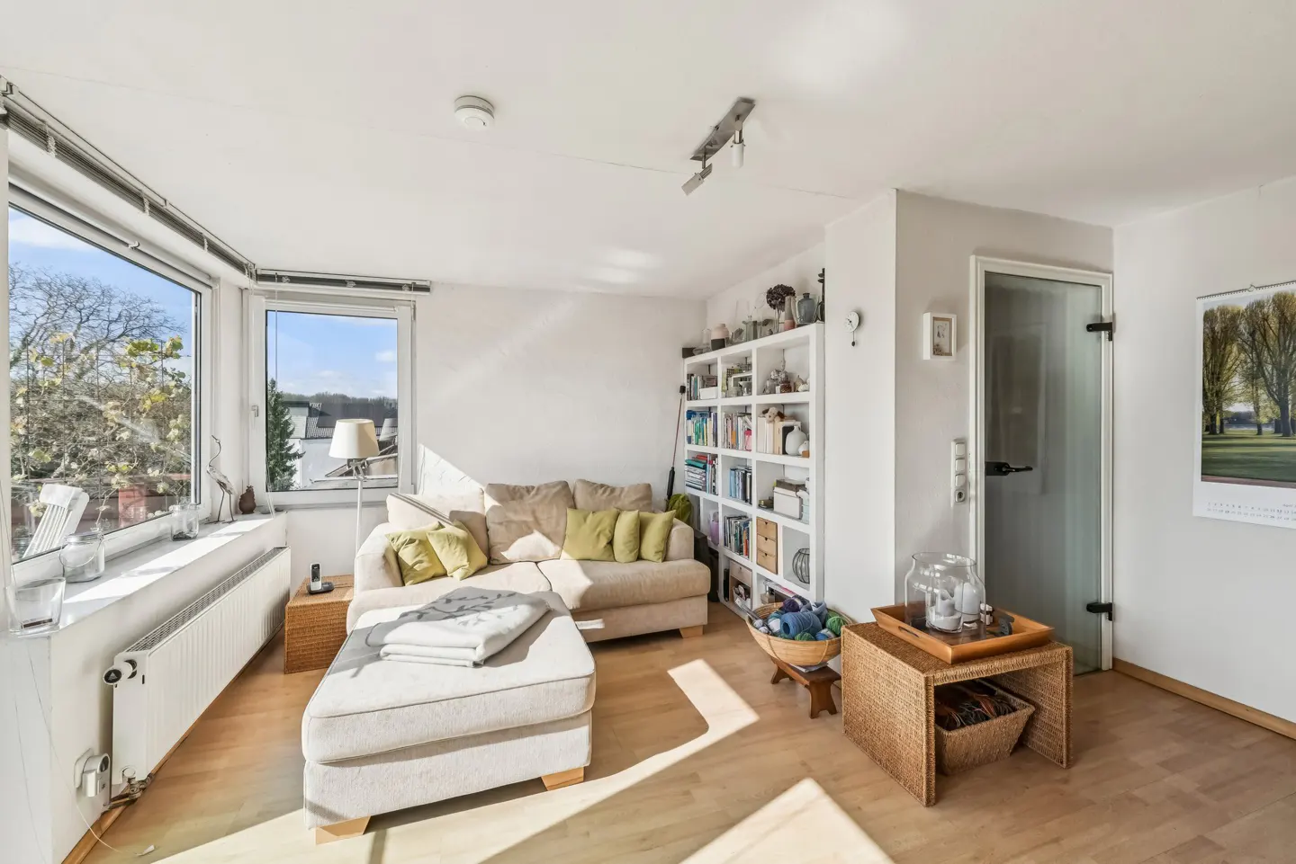 Bright living room with a beige sectional sofa, white bookcase, and large windows. Sunlight streams onto the wooden floor.