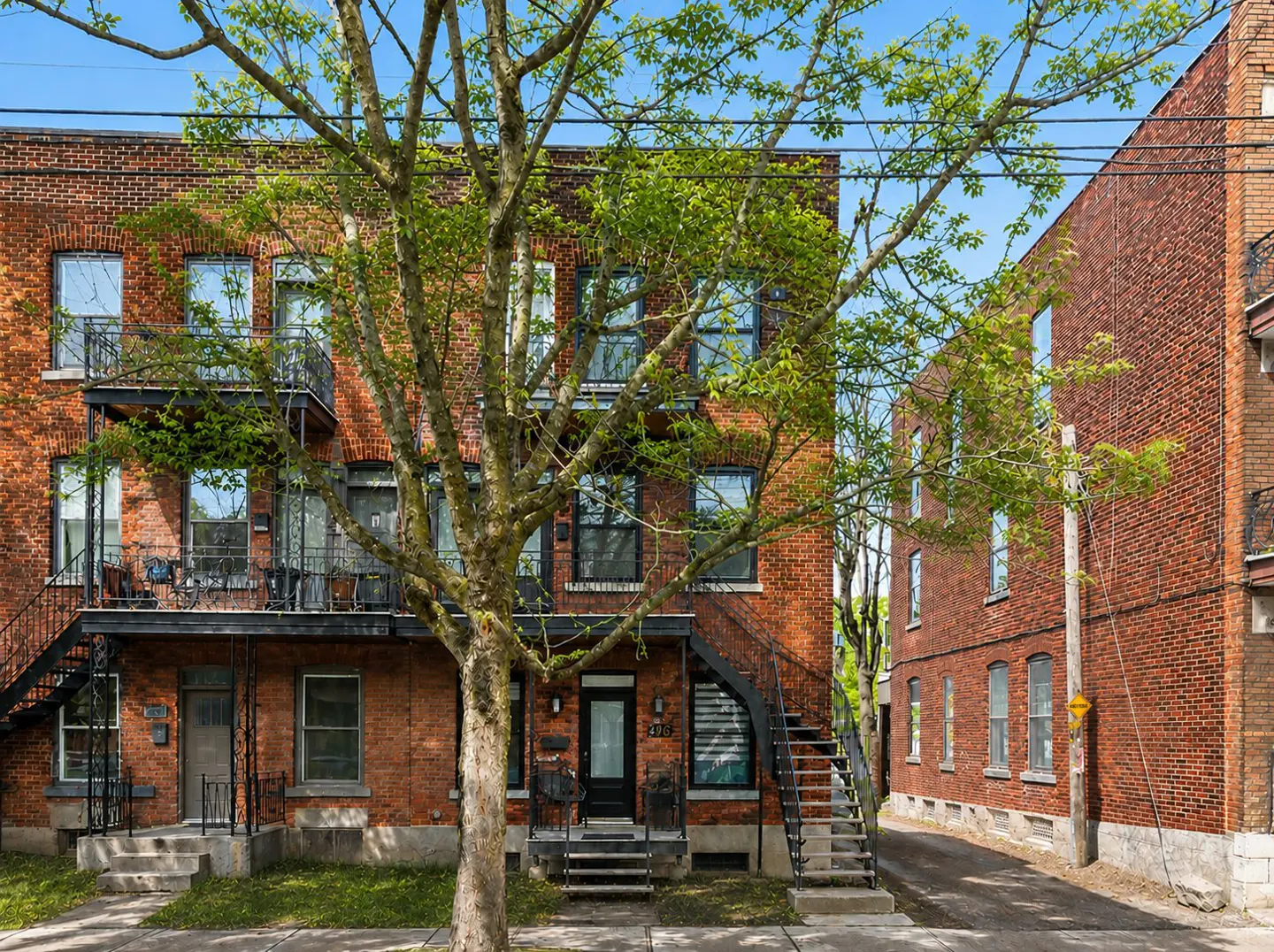 Three-story red brick apartment building with black metal balconies and exterior staircases, partially obscured by a tree.