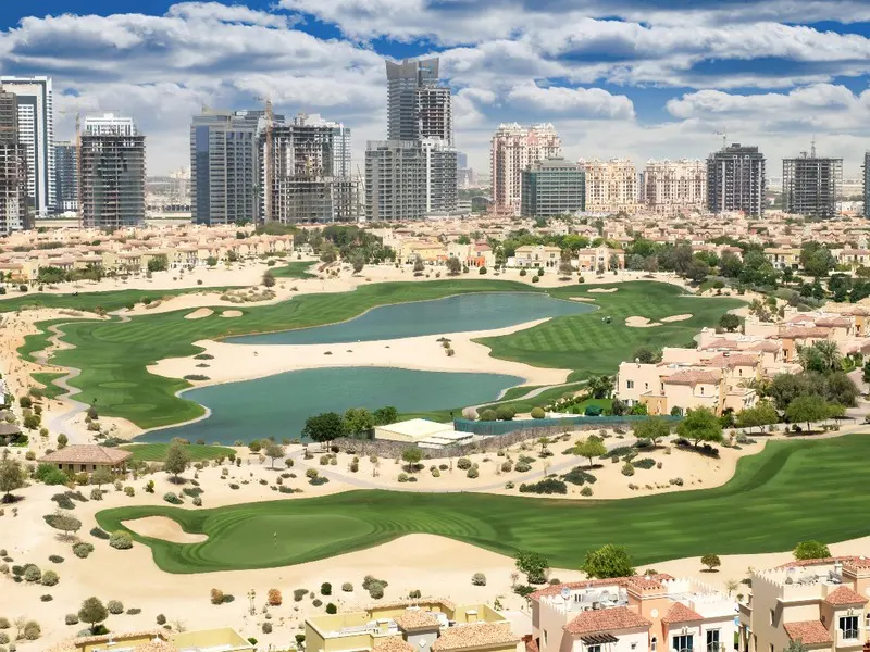 Aerial view of a green golf course with lakes, surrounded by tan desert, homes, and city skyscrapers.