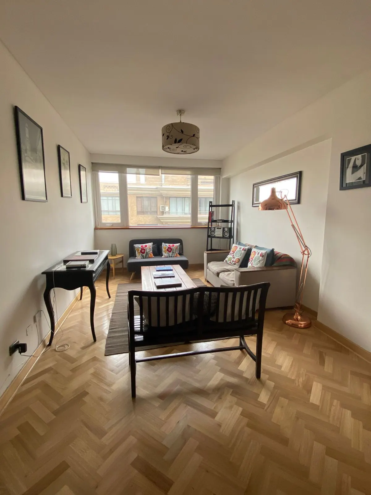 Bright living room with herringbone wood floors, sofas, and a copper floor lamp. Windows overlook a building.