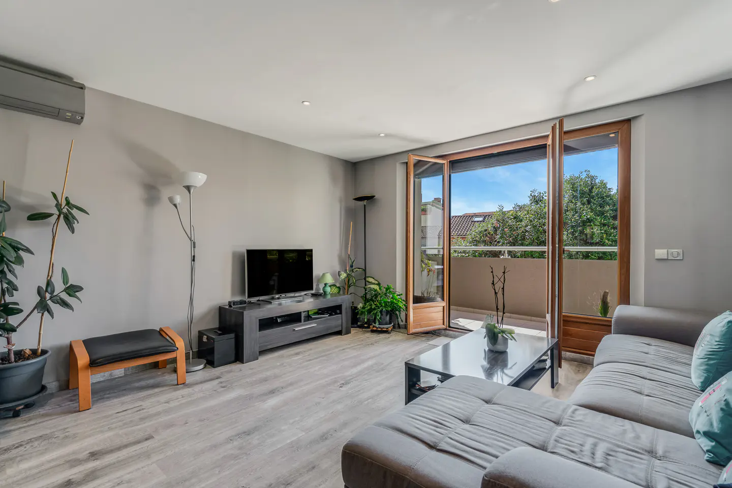 Living room with gray walls, wood floors, and a gray sectional sofa. Open doors lead to a balcony with trees and blue sky visible.
